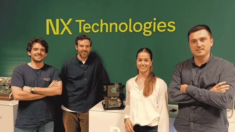 Four NX Technologies team members pose beside a hardware display beneath the company logo wall